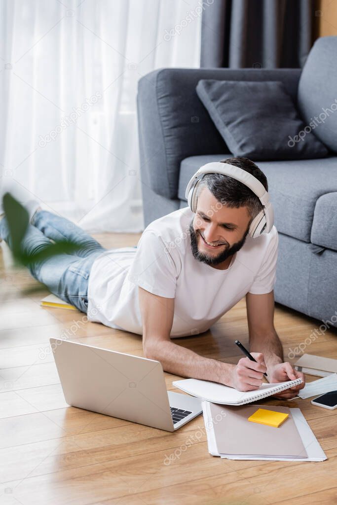 Selective focus of smiling man in headphones using laptop and stationery during online education at home
