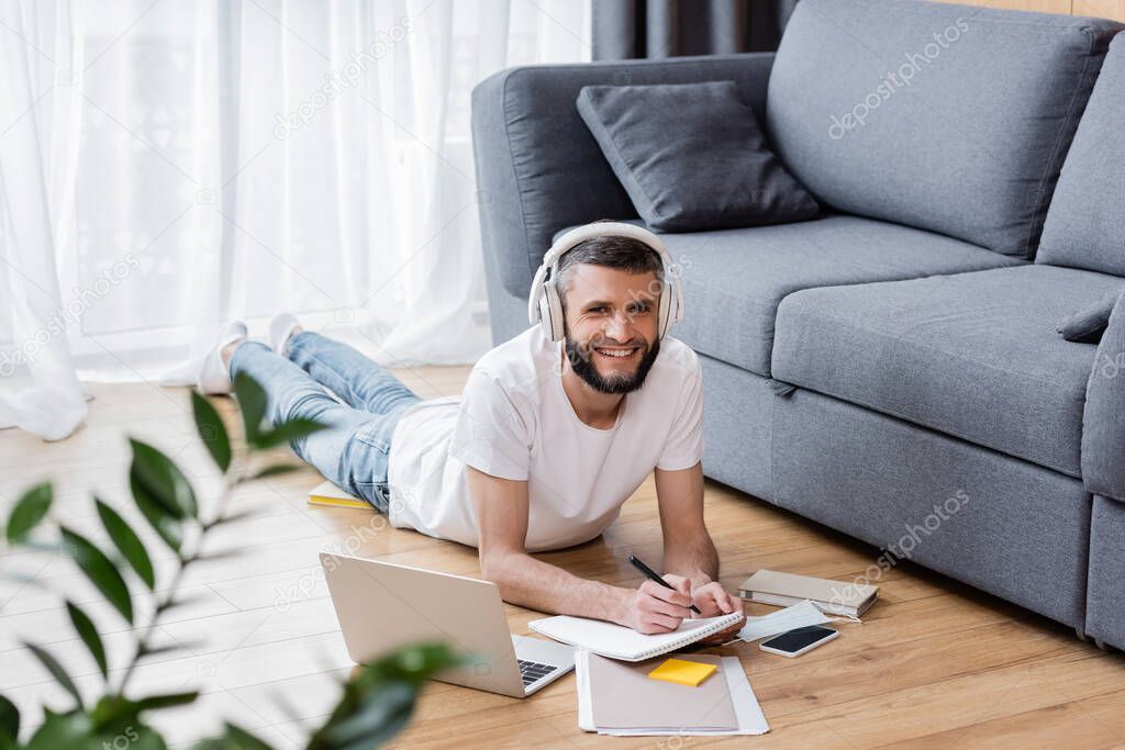 Selective focus of smiling man in headphones using stationery and laptop near medical mask on floor at home 