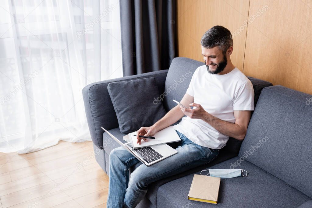 Smiling teleworker using gadgets near medical mask on couch 