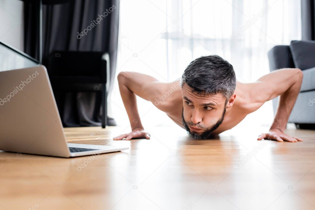 Surface level of muscular man doing push ups near laptop on floor at home 