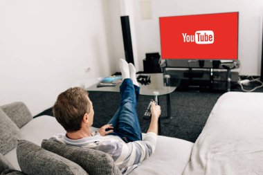 KYIV, UKRAINE - APRIL 14, 2020: man in jeans holding remote controller near tv screen with youtube at home  clipart
