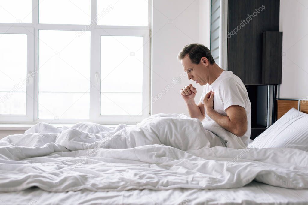 side view of sick man coughing while sitting on bed at home 