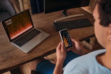 Karantina sırasında ev ofisinde grafikleri ve online ticareti olan akıllı telefon ve dizüstü bilgisayar üzerinde çalışan serbest çalışan bir erkek görülüyor.