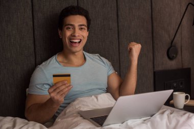 Heyecanlı karışık yarış adamı internetten kredi kartı ve laptopuyla kendini soyutlama sırasında yatakta alışveriş yapıyor.