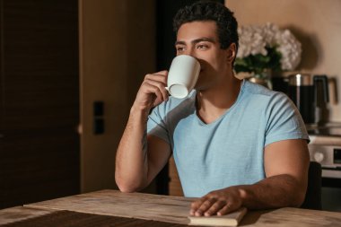 Kahve içen ve kendini izole ederken mutfakta kitap tutan melez bir adam.