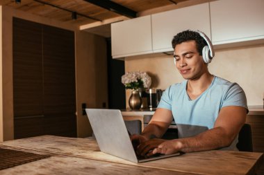 Melez adam kulaklıkla müzik dinliyor ve mutfaktaki yalnızlık sırasında dizüstü bilgisayar kullanıyor.