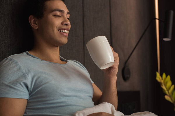 счастливый смешанной расы человек держит чашку кофе в постели на карантине
