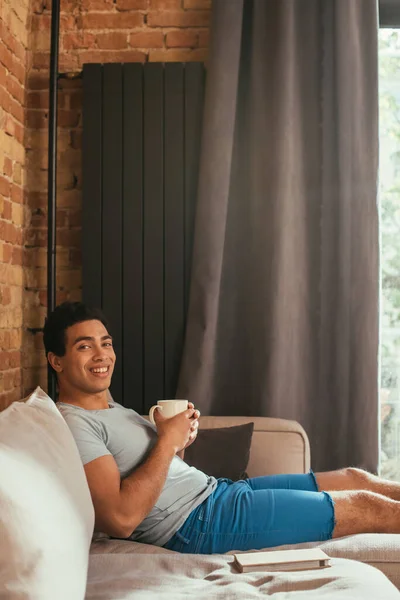 Smiling Mixed Race Man Holding Cup Coffee Sofa Book Self — Stock Photo, Image