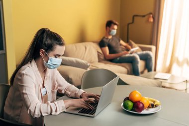 Evde dijital tabletli bir adamın yanındaki dizüstü bilgisayarı kullanarak tıbbi maskeli serbest çalışanın seçici odağı 