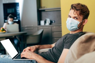 Medikal maskeli serbest çalışan kız arkadaşının yanında beyaz ekranlı dizüstü bilgisayar kullanıyor. 