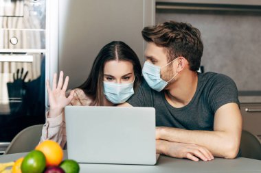 Tıbbi maskeli bir kadının, erkeğin yanında görüntülü konuşma yaparken el sallaması.