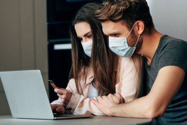 Tıbbi maskeli kadın elinde kredi kartıyla online alışveriş yapıyor. 