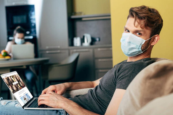КИЕВ, УКРАИНА - 22 апреля 2020 года: избирательный фокус фрилансера в медицинской маске с помощью ноутбука с веб-сайта depositphotos рядом с подругой дома
