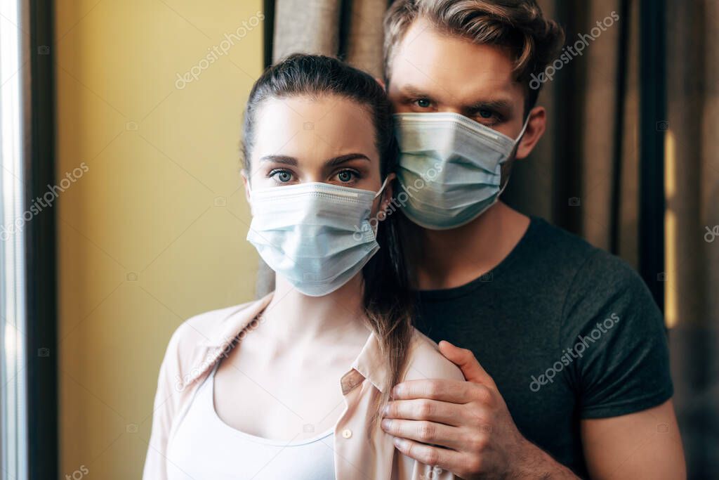 man hugging girlfriend in medical mask and looking at camera 