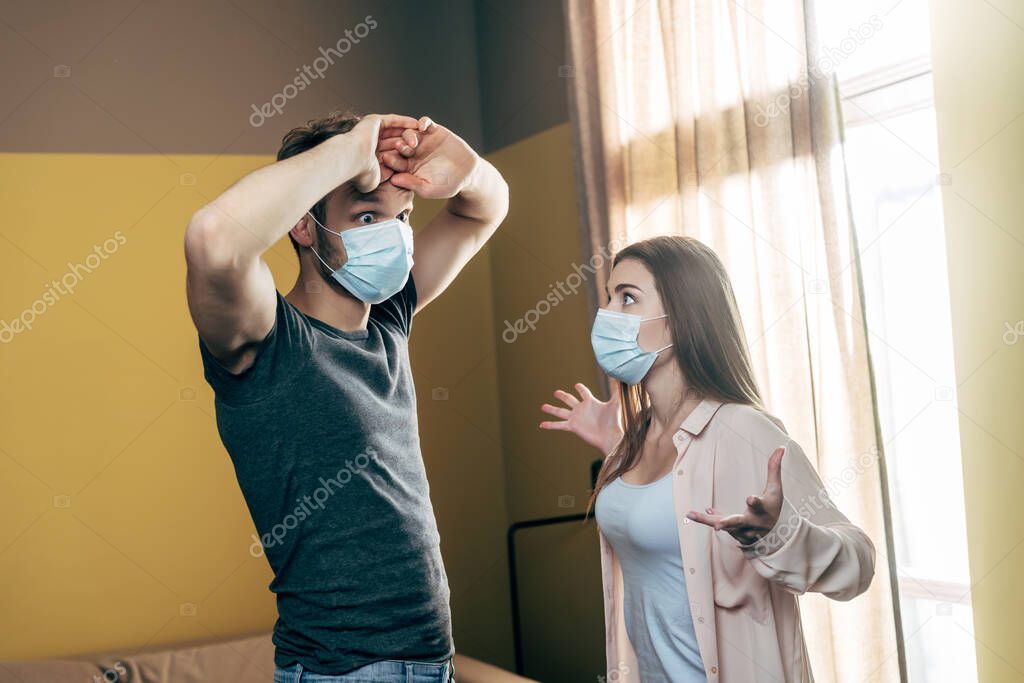 woman in medical mask gesturing while quarreling with boyfriend at home