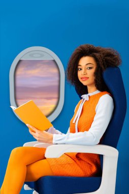 smiling african american woman in retro dress holding book and sitting on seat isolated on blue with porthole clipart