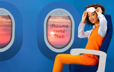 Portakal rengi retro elbiseli gülümseyen Afrikalı Amerikalı kadın elinde uyku maskesiyle mavi arka planda oturuyor ve pencereler ve hayaller gerçek oluyor.