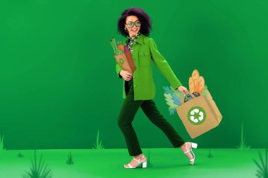 Gülümseyen ve şık Afrikalı Amerikalı kadın yeşil arka planda geri dönüşüm işaretleri olan resimli alışveriş torbalarıyla yürüyor. 
