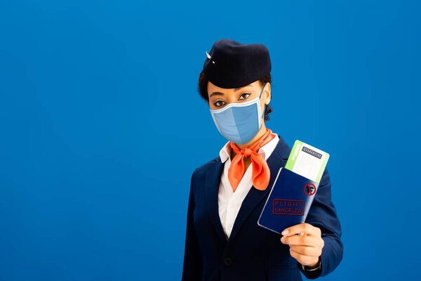african american flight attendant in medical mask holding passport and air ticket isolated on blue 