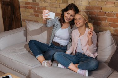 Annesinin yanında akıllı telefonuyla selfie çekerken gülümseyen çocuk barış işareti gösteriyor. 