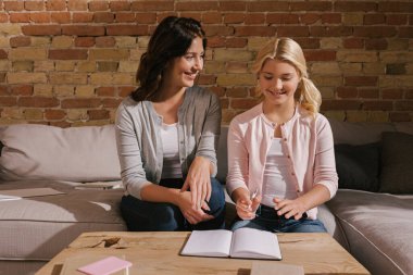 Gülümseyen kadın, oturma odasındaki sehpanın üzerinde elinde kalem ve kitaplar tutan kızına bakıyor.