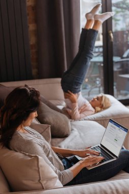 KYIV, UKRAINE - 24 Nisan 2020: Dizüstü bilgisayarlı bir kadının kanepenin yanındaki kızının yanında facebook web sitesi kullanmasının seçici odağı 