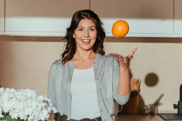 Foco Seletivo Mulher Sorridente Jogando Laranja Perto Flores Cozinha — Fotografia de Stock