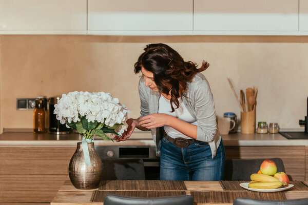 Attractive woman touching flowers in vase near fruits on kitchen table 