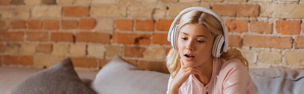 Panoramic shot of dreamy kid in headphones sitting on couch 