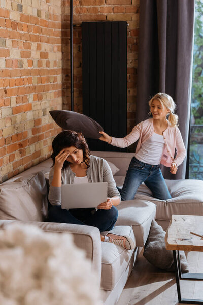 Selective focus of cheerful kid playing pillow fight with mother working on laptop at home 