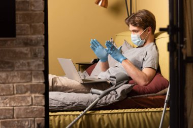 Tıbbi maskeli ve lateks eldivenli engelli bir adamın yatak odasındaki laptopta görüntülü konuşma yapması.