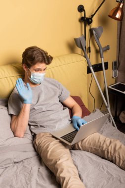 Tıbbi maskeli engelli bir adamın yatak odasındaki koltuk değneklerinin yanında dizüstü bilgisayarında görüntüsü var.