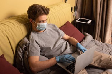 Tıbbi maskeli ve lateks eldivenli bir adam laptopla yatakta çalışıyor. 
