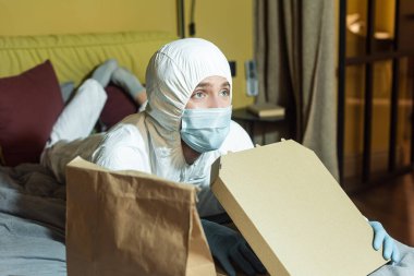 Tıbbi maskeli ve tehlikeli madde giysili bir adamın yatakta paketin ve pizza kutusunun yanında televizyon izlemesinin seçici odağı. 