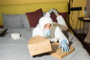Tehlikeli madde giysisi ve tıbbi maskeli bir adam paketler ve yatağın üzerinde pizza kutusunun yanında yatıyor. 