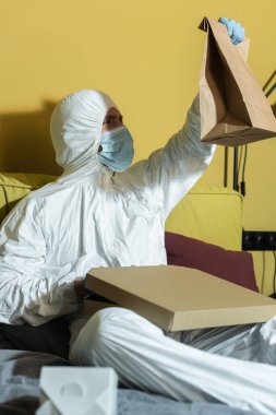 Tıbbi maskeli ve tehlikeli madde giysili bir adamın seçmeli odak noktası yatağın yanındaki pizza kutusunun yanındaki pakete bakması. 
