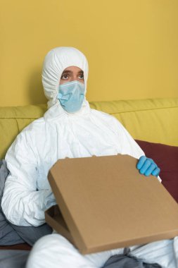 Tıbbi maskeli ve tehlikeli madde giysili bir adamın yatakta pizza kutusunu tutmasının seçici odağı. 
