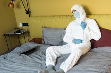 Tehlikeli madde giysisi, tıbbi maske ve lateks eldivenli bir adam yatakta dizüstü bilgisayarın yanında oturuyor. 