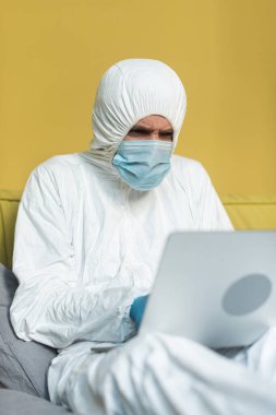 Tehlikeli madde giysisi ve tıbbi maskeli bir adamın yatakta dizüstü bilgisayar kullanmasının seçici odağı. 