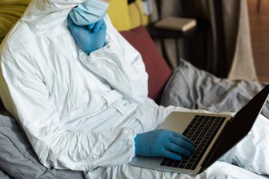 Kanepede dizüstü bilgisayar kullanan tehlikeli madde giysisi ve tıbbi maske takmış bir adamın görüntüsü. 