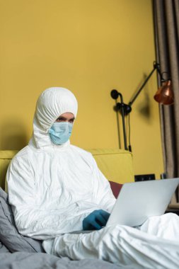 Tehlikeli madde giysisi ve tıbbi maskeli bir adamın yatakta dizüstü bilgisayarda çalışmasının seçici odağı. 