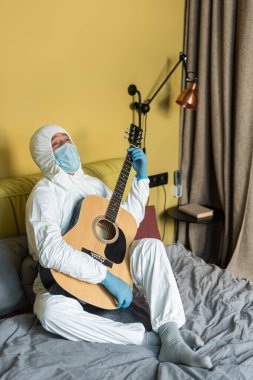 Tehlikeli madde kıyafeti giymiş bir adam, tıbbi maske ve akustik gitar tutan lateks eldivenler. 