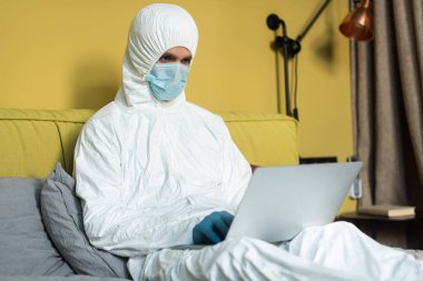 Yatak odasında dizüstü bilgisayar kullanan tehlikeli madde kıyafeti giyen ve tıbbi maske takan bir adamın seçici odağı. 