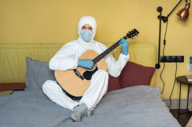 KYIV, UKRAINE - 24 Nisan 2020: Tehlikeli madde giysisi ve tıbbi maskeli bir adam akustik gitar tutuyor ve kanepede kameraya bakıyor 