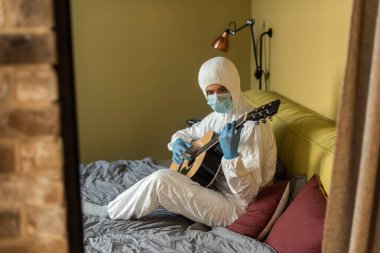 Tehlikeli madde giysisi ve lateks eldivenli bir adamın yatak odasında akustik gitar çalmasının seçici odağı.