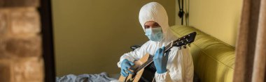 Tehlikeli madde giysisi ve tıbbi maske takmış yatak odasında gitar çalan panoramik bir adam.