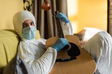 Tehlikeli madde giysisi ve tıbbi maskeli bir adamın yatakta akustik gitar çalarken kameraya baktığı seçici odak noktası. 