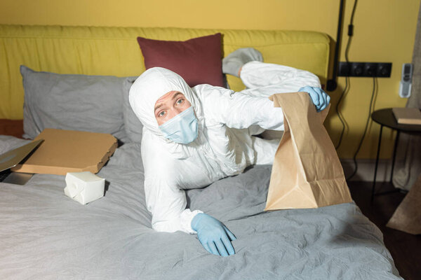 Man in hazmat suit holding package while lying near laptop and pizza box on bed 
