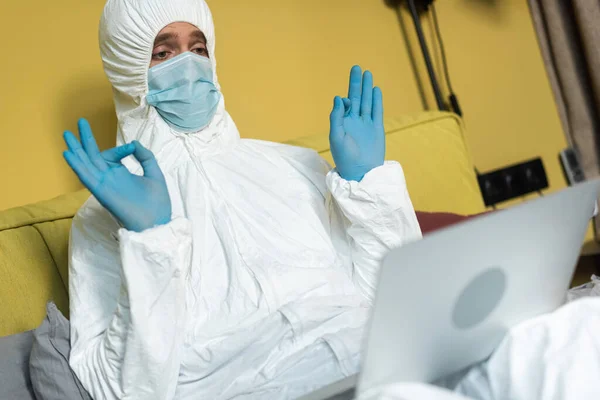 Selective focus of man in medical mask and hazmat suit showing okay sign during video call on laptop on bed