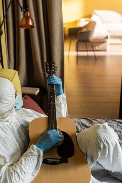 穿着危险物品套装 医疗面罩和乳胶手套在床上弹奏吉他的男子侧视图 — 图库照片